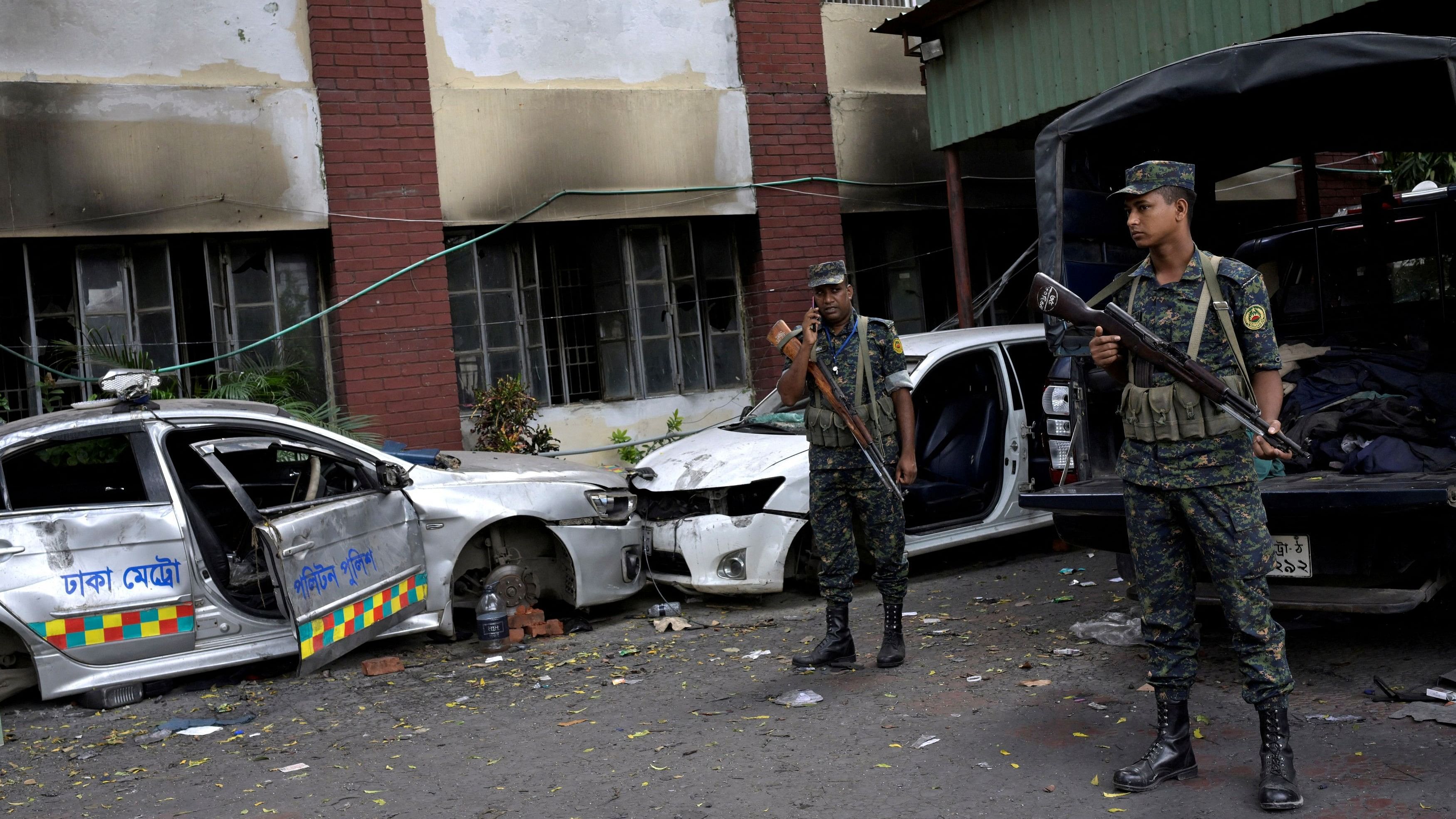 <div class="paragraphs"><p>Security force personnel stand guard next to damaged vehicles outside a police station, in Dhaka</p></div>